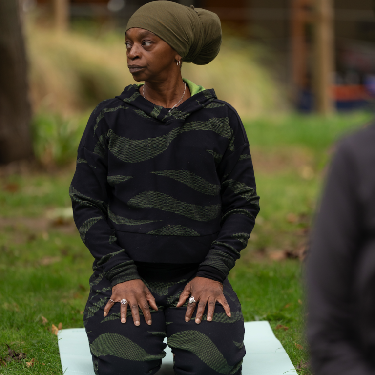 Fiery Hearts festival female attendee in a camouflaged outfit sits on a mat in a park, engaged in yoga.