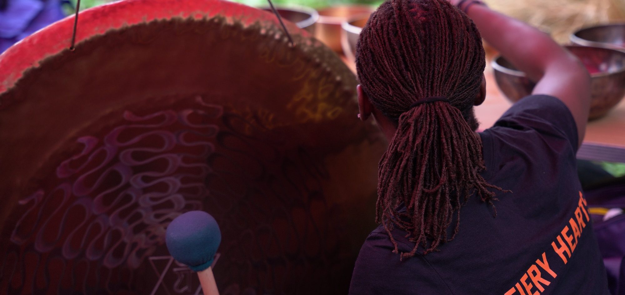 Person playing a large gong viewed from behind, with long hair and a black Fiery Hearts T-shirt.